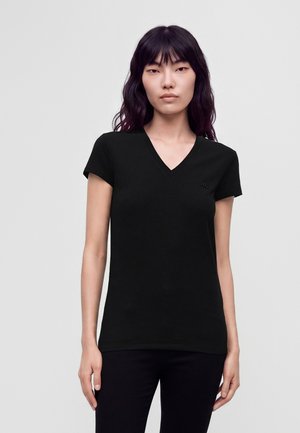 Jeune femme aux longs cheveux foncés portant un t-shirt noir ajusté à col en V et manches courtes sur un fond clair uni.