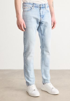 Man draagt lichtblauwe slim-fit jeans met witte sneakers, staand tegen een effen achtergrond.