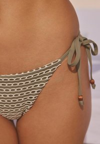 Teksturirani bikini donji dio u maslinastoj zelenoj boji s uzorkom kružnih izreza. veze sa strane sadrže drvene perle kao detalj.