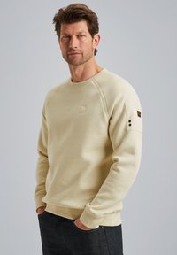 Sweat-shirt beige avec un col rond et des manches raglan, arborant un logo en patch sur le devant et un détail de bouton sur la manche gauche. Tissu doux.