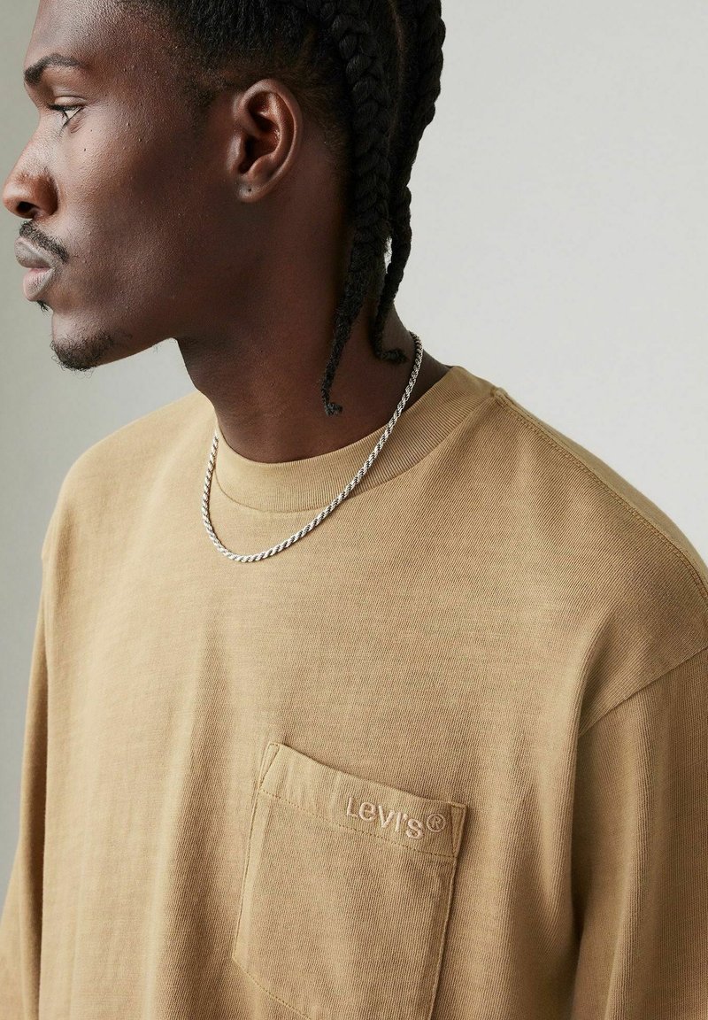 Camisa de manga larga beige con un bolsillo en el pecho izquierdo que presenta un logo bordado. Un collar de cadena plateada completa el look.