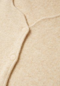 Zbliżenie beżowego, miękkiego kardiganu z dzianiny z dwiema widocznymi, dopasowanymi guzikami oraz widocznym szwem wzdłuż linii guzików.