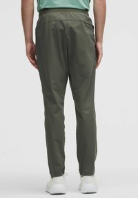 Person klädd i olivgröna slim-fit byxor, vita sneakers och en instoppad ljusgrön skjorta, stående med händerna längs sidorna, vänd bort.