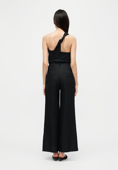 Μαύρη ολόσωμη φόρμα με έναν ώμο, με λεπτομέρεια δέσιμου στον ώμο. Διαθέτει παντελόνι με φαρδιά πόδια και απαλό υλικό.