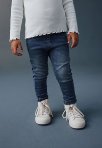 Fehér bordázott hosszú ujjú ing fodros szegéllyel, sötétkék szűk farmernadrággal és fehér, fűzős magas szárú tornacipővel párosítva.