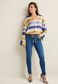 Blouse tie-dye multicolore dans des tons de brun, bleu et crème, avec des manches larges et une taille à cordon, associée à un jean bleu ajusté.