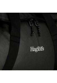 Borsa da viaggio nera realizzata in materiale resistente, con due manici robusti, chiusura con zip e logo "Haglöfs" bianco. Superficie testurizzata.