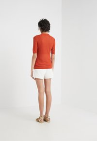 Person med kort, krøllet hår står med ryggen til, iført en struktureret orange T-shirt med korte ærmer, hvide shorts og guld sandaler.