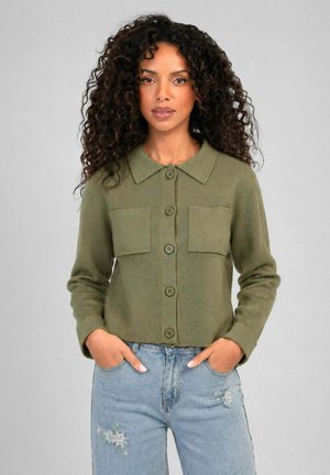 Femme aux cheveux bouclés portant une chemise boutonnée vert olive avec des poches à l'avant et un jean bleu clair avec une broderie florale, mains dans les poches.