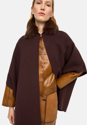 Kobieta ubrana w brązowy płaszcz z futrzanym kołnierzem, na skórzanej, beżowej kurtce i spódnicy, pozująca z jedną dłonią na biodrze na jednolitym jasnym tle.