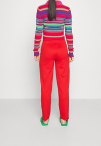 Femme aux longs cheveux noirs portant un haut à rayures colorées à manches longues, un pantalon rouge et des chaussures vertes à enfiler, se tenant de dos.