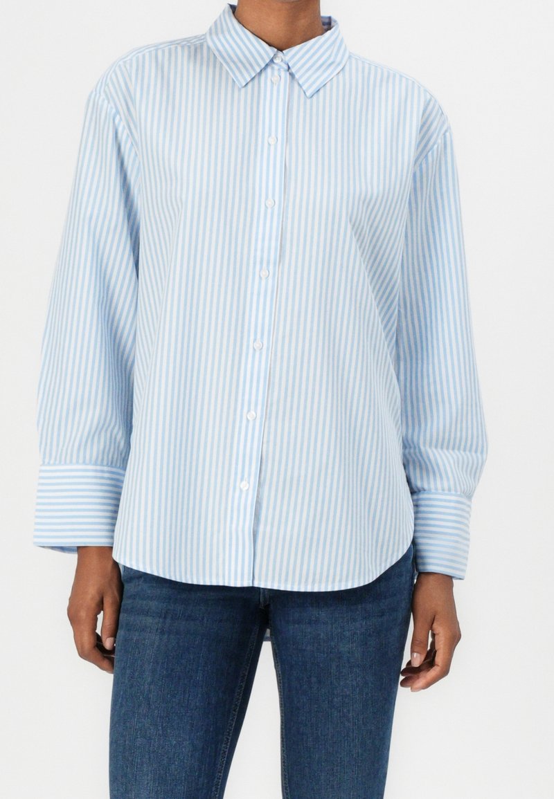 Personne portant une chemise à rayures verticales bleu clair et blanc à boutons, à manches longues, et un jean bleu foncé.