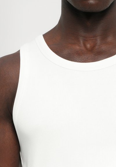 Débardeur blanc à côtes avec un col rond, épousant étroitement les épaules et la poitrine, mettant en valeur une texture lisse.