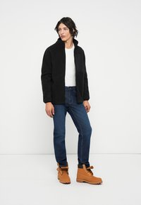 Svart fleecejakke med høy krage; hvit t-skjorte under. Blå jeans og lysebrune snørestøvler med svarte detaljer. Uformelt antrekk.