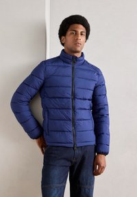 Navyblaue Daunenjacke mit hohem Kragen, horizontal gestepptem Design, Frontreißverschluss und Seitentaschen. Strukturierten Stoff und elastische Bündchen.