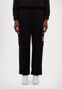 Zwarte wijde sweatpants gemaakt van zachte stof met zijzakken; heeft een witte logo-detail op de linkerzak en is gecombineerd met witte sneakers.