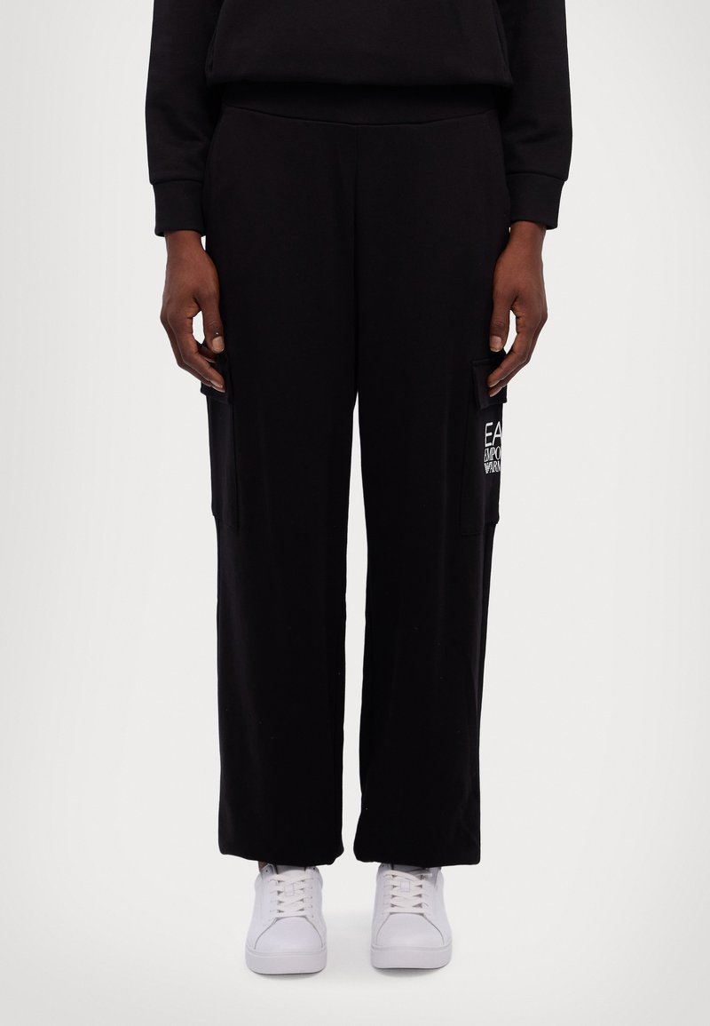 Zwarte wijde sweatpants gemaakt van zachte stof met zijzakken; heeft een witte logo-detail op de linkerzak en is gecombineerd met witte sneakers.