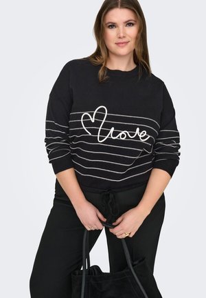 Kobieta w czarnym swetrze z białym napisem "love" i sercem, czarnych spodniach, trzymająca czarną torebkę, pozująca na jednolitym tle.