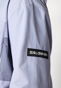 Hele lilla jakk, millel on tekstureeritud viimistlus, must "SALOMON" logo padi varrukal ning kanga peen ruuduline muster.