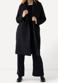Cappotto in lana nera con design doppiopetto, ampi revers, tasche laterali e vestibilità comoda, indossato sopra pantaloni e scarpe scure.