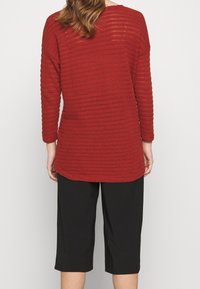 Tunique rossa a maniche lunghe e costine con orlo arrotondato, abbinata a pantaloni neri a tre quarti. Texture di maglia leggera visibile nel tessuto.