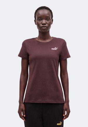 Γυναίκα που φοράει ένα απλό σκούρο βυσσινί t-shirt Puma με λογότυπο στο στήθος, στέκεται ακίνητη προς τα εμπρός με φόντο ένα απλό ανοιχτό χρώμα.