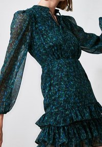 Blommig klänning i mörkgrön med blå detaljer. Har en volangnedre kant, långa genomskinliga ärmar och knäppning vid halsen. Lättviktsmaterial.