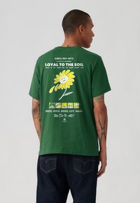 Tricou verde din bumbac cu un grafic de margaretă galbenă și text pe spate, mâneci scurte, croială lejeră, având detalii imprimate și guler rotund.