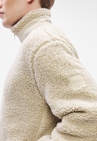 Bež flis pulover s visokim ovratnikom i teksturiranom površinom. Na rukavu ima mali brandirani zakrpu. Glatki materijal ispod ovratnika.