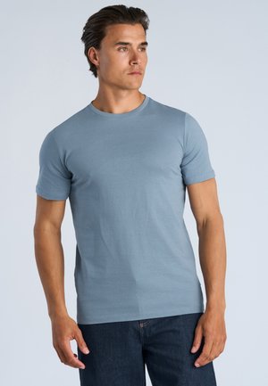 Mann mit dunklen Haaren, der ein hellblaues kurzärmeliges T-Shirt und dunkle Jeans trägt und leicht nach rechts schaut vor einem einfarbigen Hintergrund.
