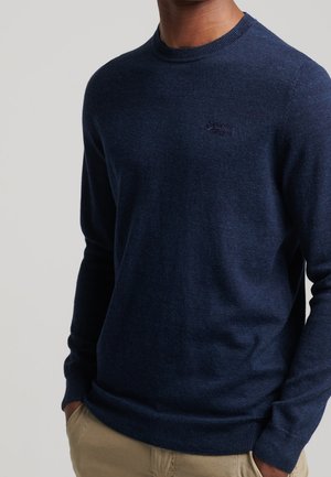 Man draagt een marineblauwe trui met ronde hals en subtiel geborduurd logo op de borst, en beige broek, handen in de zakken.