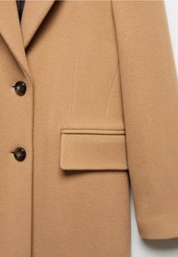 Manteau en laine couleur fauve avec deux boutons foncés et une poche à rabat rectangulaire sur le côté droit, montrant la partie supérieure avant et une partie de la manche.