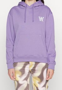 Fioletowy hoodie z przednią kieszenią, kapturem ze sznurkiem oraz białym logo na klatce piersiowej. Miękki materiał i luźny krój. Połączony z wzorzystymi spodniami.