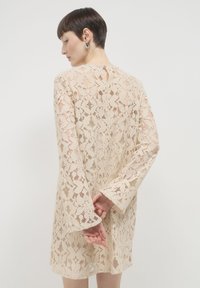 Beiges Spitzenkleid mit langen Glockenärmeln und floralem Muster. Schlüssellochdetail am Rücken und lockere Passform. Weicher, strukturierter Stoff.