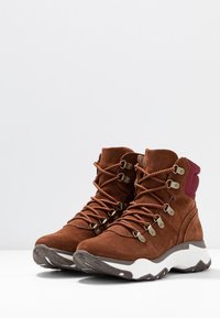 Brązowe zamszowe buty turystyczne z wysokim designem kostki, z metalowymi oczkami, okrągłymi sznurowadłami oraz białą podeszwą pośrednią i wzorzystą brązową podeszwą zewnętrzną.