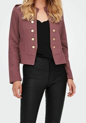 Mujer con chaqueta corta malva con botones dorados sobre un top negro y pantalones negros acolchados de talle alto sobre un fondo blanco.