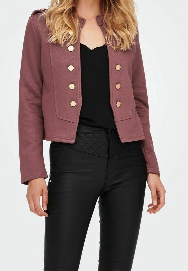 Femme portant une veste courte mauve avec des boutons dorés sur un top noir et un pantalon noir matelassé taille haute sur fond blanc.
