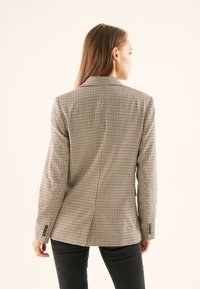 Kvinde med langt hår iført en brun og hvid ternet blazer og sorte bukser, set bagfra mod en ensfarvet lys baggrund.