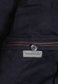 Sötét szövet sima textúrával, barna bőr cipzárdíszítéssel, gombos záródással és egy címkével, amelyen az szerepel: "Till Death Do Us Part" (Míg a halál el nem választ).