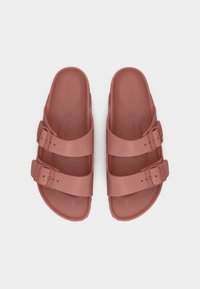 Ζευγάρι σαγιονάρες σε χρώμα σκόνης ροζ με δύο ρυθμιζόμενους ιμάντες με αγκράφες, επίπεδες σόλες και ανάγλυφα λογότυπα στη σόλα, σε άποψη από πάνω.