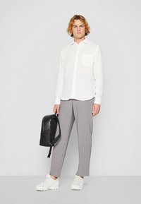 Weißes Button-Up-Hemd mit einer einzelnen Brusttasche, kombiniert mit grauen Nadelstreifenhosen. Schwarzer Rucksack und weiße Sportschuhe inklusive.