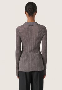 Suéter marrón de manga larga con textura acanalada y una silueta ajustada, acompañado de un cuello amplio. La tela tiene una textura ligeramente brillante. Combinado con pantalones negros.