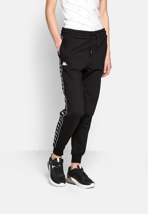 Pantaloni jogging neri con elastico in vita, tasche laterali e logo bianco. Presentano strisce laterali a fantasia contrastante. Abbinati a sneakers nere.