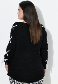 Frau mit langen glatten Haaren, die einen schwarzen Pullover mit weißen Schleifen an den Ärmeln trägt, steht vor einem schlichten weißen Hintergrund.