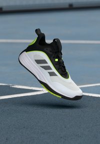 Weißer, schwarzer und neon-grüner Sportschuh schwebt in der Luft über einem blauen Tennisplatz mit weißen Linien, aus seitlicher Perspektive gesehen.