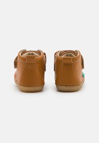 Chaussures pour enfants en cuir brun avec des bandes Velcro, des talons arrondis et des accents cousus, comportant une petite étiquette verte sur le côté.