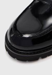 Schwarzer, glänzender Lederschuh mit detaillierter Naht und dicker, schwarz strukturiertem Gummisohle auf hellgrauem Hintergrund.