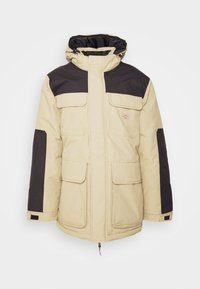 Beige, polstret jakke med sorte detaljer, med høyt krage, flere frontlommer og glidelås. Strukturert stoff for varme.