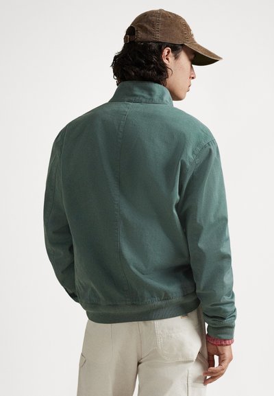 Πράσινο καμβά μπουφάν με γιακά, ριμπ μανσέτες και ζώνη στη μέση. Η πλάτη έχει απλό σχέδιο χωρίς ορατά λογότυπα ή μοτίβα.