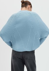 Donna con i capelli raccolti in un bun, che indossa un maglione oversized a coste di colore azzurro chiaro e jeans scuri, vista di spalle su uno sfondo semplice.